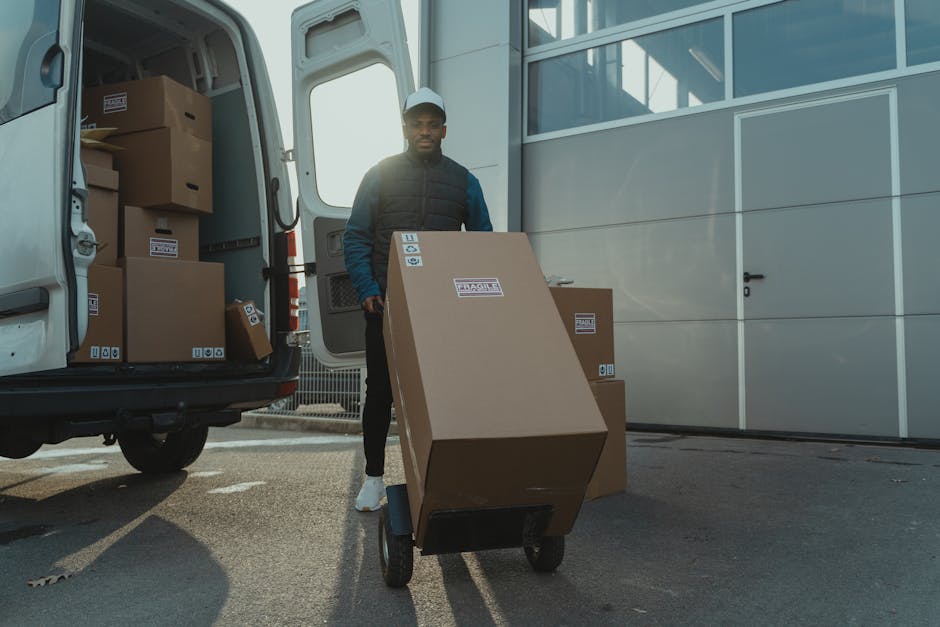 A young male mover with long, wavy hair wearing a patterned headband and a dark shirt is sitting in the driver’s seat of a white moving van with the company's branding, including the words 'MOVING C