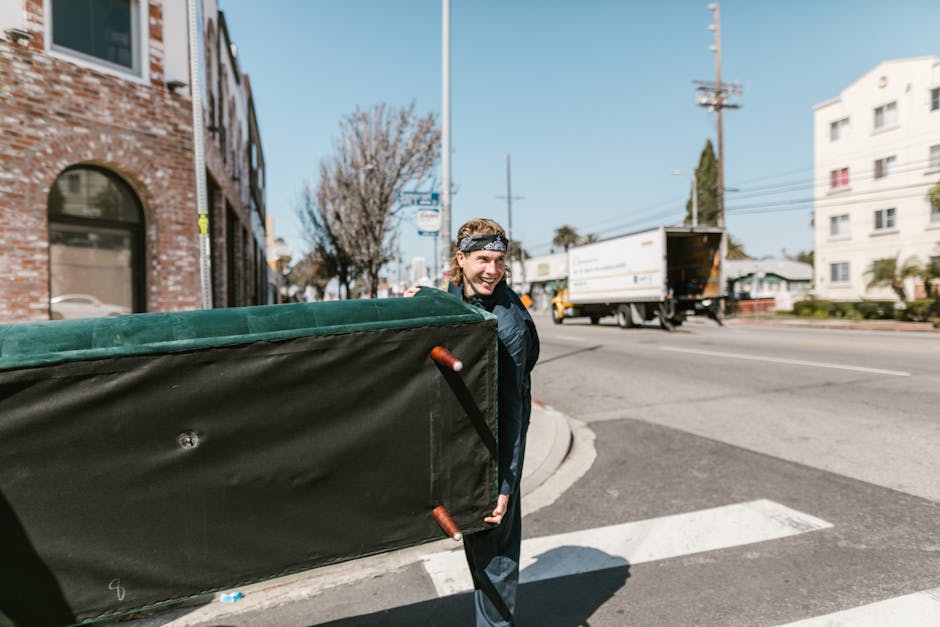 A person wearing a dark green t-shirt and blue work trousers is in the process of moving a cardboard box, which has a white and red safety label that reads 'Caution This Side Up' with an upward arrow,