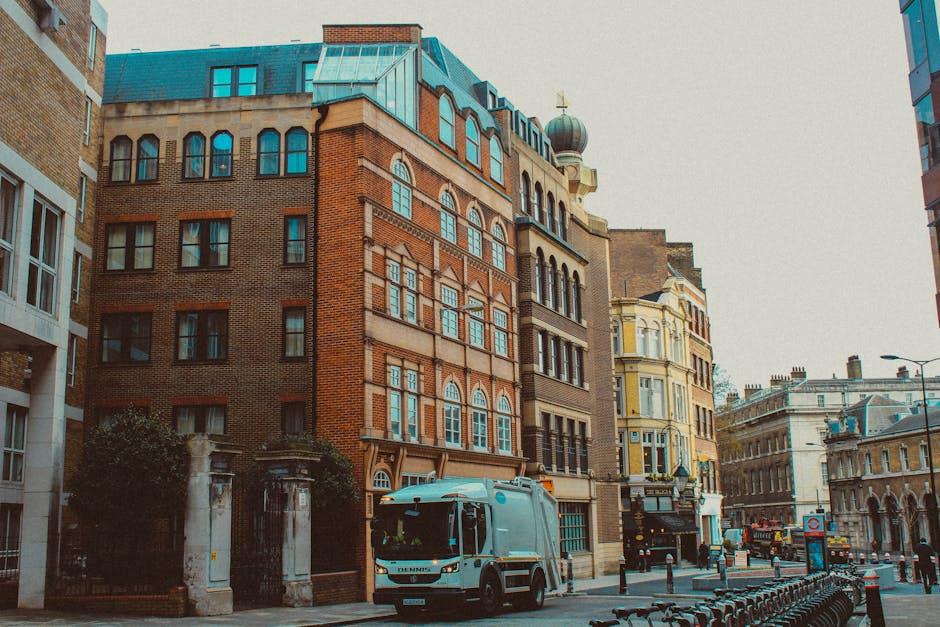 A mid-sized white and blue van belonging to Clapham Removals is parked on a city street in front of a row of multi-storey buildings with varied architectural styles, including brick and ornate facades